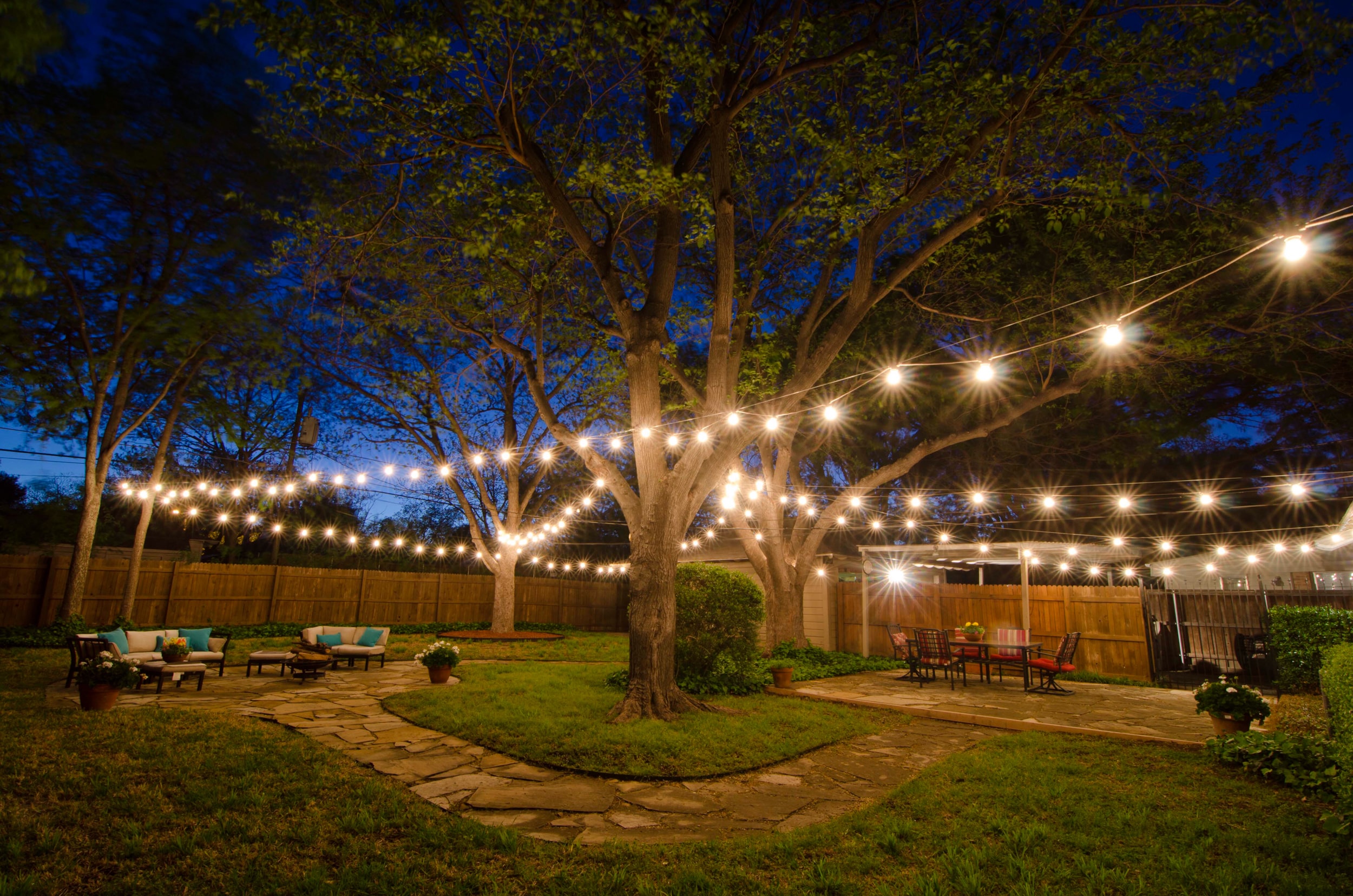 A serene backyard illuminated by string lights, creating a warm and inviting atmosphere. The soft glow of the lights adds a magical touch to the outdoor space, casting a gentle radiance on the surrounding plants and furniture. The strings of lights are delicately draped across the yard, forming a mesmerizing pattern against the night sky. The cozy ambiance invites relaxation and outdoor gatherings, making it a perfect setting for socializing or enjoying a peaceful evening under the stars.
